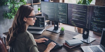 A woman in glasses coding on multiple computer screens.